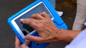A hand presses an icon on the screen of a blue Seedling educational touchpad.